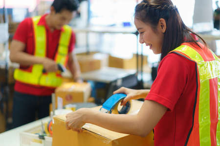Asian female worker packing cardboard box with tape gun dispenser in warehouse. Thai employee packing goods in large industrial storehouse.の写真素材
