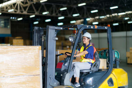 Asian beautiful woman driver drive fork lift car in industry with smile, ability of girl and diversity of career wearing helmet in reflective vest working on vehicleの写真素材
