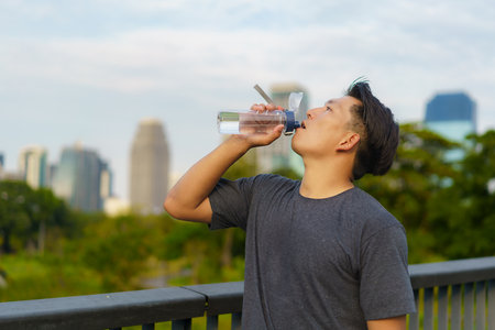 Image of Asian healthy man holding a bottle of refreshing water, resting after a workout or a run in city parkの写真素材