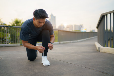 Close-up of Asian man tying shoelaces on white sneakers. Male person squatting on sidewalk. Sunny morning or day. Male going on footpath. Sport and healthy lifestyle conceptの写真素材