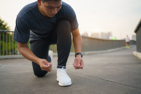 Close-up of Asian man tying shoelaces on white sneakers. Male person squatting on sidewalk. Sunny morning or day. Male going on footpath. Sport and healthy lifestyle conceptの写真素材