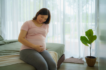 Pregnancy, people rest and expectation concept - Asian smiling happy pregnant woman sitting in bed and touching her belly at home.の写真素材