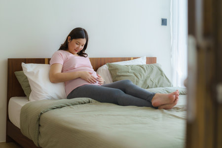 Pregnancy, people rest and expectation concept - Asian smiling happy pregnant woman sitting in bed and touching her belly at home.の写真素材