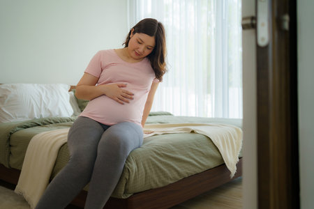 Pregnancy, people rest and expectation concept - Asian smiling happy pregnant woman sitting in bed and touching her belly at home.の写真素材