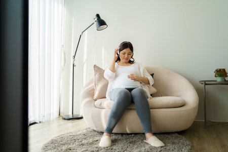 Portrait of pregnant Happy Asian woman sitting on sofa at home and using smart phone for listening music in headphones.の写真素材