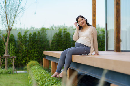 Portrait of pregnant Happy Asian woman sitting on terrace at home and using smart phone for listening music in headphones.の写真素材