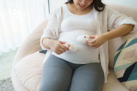 Pregnant woman holding baby booties and sitting on sofa at homeの写真素材