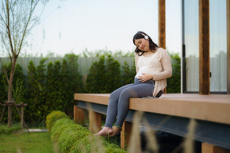 Pregnant woman listening to music on headphones sitting on terraceの写真素材