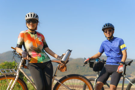 Tired Asian cyclist couple taking a break to drink water from a bottle with a beautiful lake and mountains in the background.の写真素材
