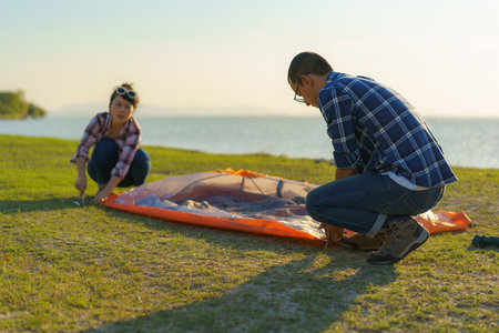 Asian couple preparing a tent to camping in the lawn with the lake in the background during sunsetの写真素材