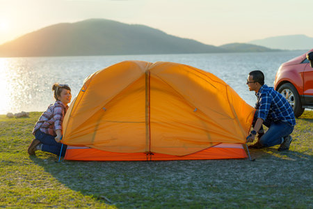 Asian couple preparing a tent to camping in the lawn with the lake in the background during sunsetの写真素材