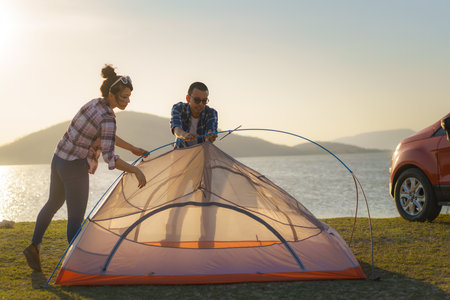 Asian couple preparing a tent to camping in the lawn with the lake in the background during sunsetの写真素材