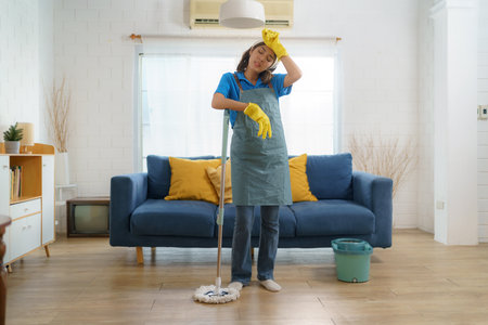 Exhausted Asian maid struggles with fatigue while mopping the house, showcasing the challenges and hard work behind domestic houseの写真素材