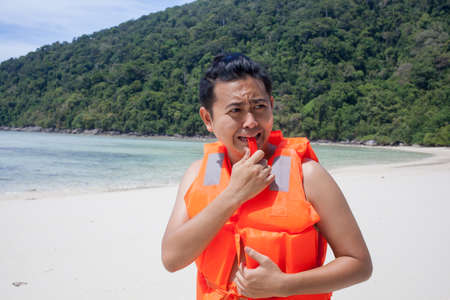 lifeguard, a male model wearing light orange life jacket with a whistle standing on the beach acting funny by the seaの写真素材