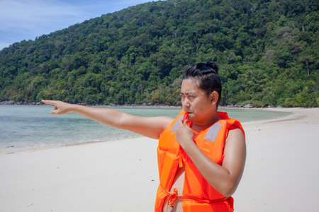 lifeguard, a male model wearing light orange life jacket with a whistle standing on the beach acting funny by the seaの写真素材