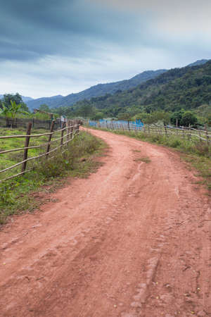 pathway, red dirt rough pathway road along bamboo fence lead to farm with mountain view, green environment and skyの写真素材