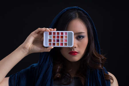 A girl wearing  make-up in mysterious look holding mix color lipstick pad on black backgroundの写真素材