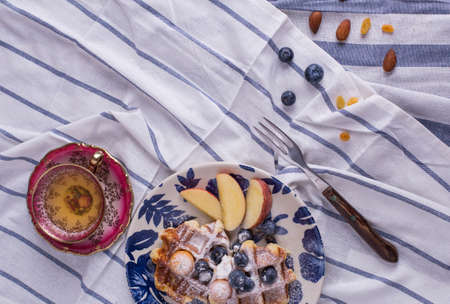 Blueberry apple waffle with honey set on a dish decorated with fresh blueberry, apple, tea pot with copy space.の写真素材