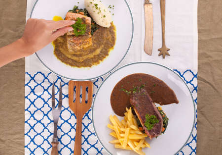 A hand decorating nice dish of salmon steak and beef served with mashed potato,lemon, saute spinach and sauce on background with wooden utensils.の写真素材