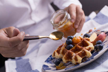 Blueberry apple waffle with honey set on a dish with a chef's hand holding spoon for a jar of honey ready to serve.の写真素材