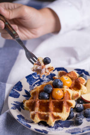 Closeup of a fork cutting a piece of blueberry waffle decorated with fresh blueberry in a nice dish.の写真素材