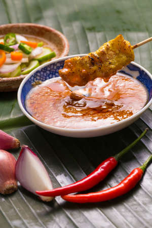 A hand having pork satay dipping peanut sauce decorated with cucumber sauce, onion, chili on banana leaf.の写真素材
