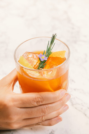 Orange color cocktail in a rocks glass decorated with orange peel, rosemary and purple flower in a...