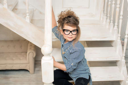 Beautiful young girl in glasses sitting on the stairs of the bright roomの写真素材