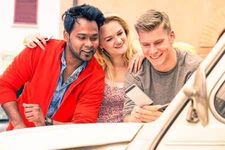 Multiracial young friends watching mobile phone on car bonnet - Cheerful guys leaning on vintage auto using smartphone technology - Concept of multicultural friendship and joyful moment on a road tripの写真素材