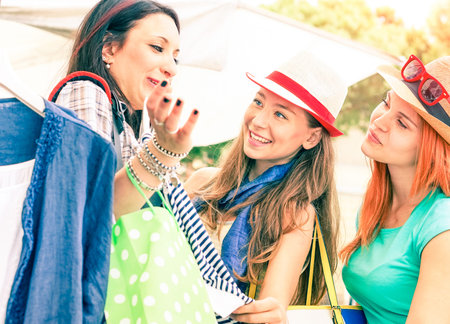 Travel girlfriends at flea market fun and shopping moment - Young happy girls checking shirts size at street shop on summer vacation day - Cheerful women talking together - Focus on middle femaleの写真素材