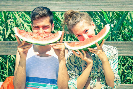Positive couple eating watermelon at countryside picnic - Young friends having fun outdoor playing funny faces with seasonal red fruit - Concept of joyful moment on summertime - Vintage filter lookの写真素材