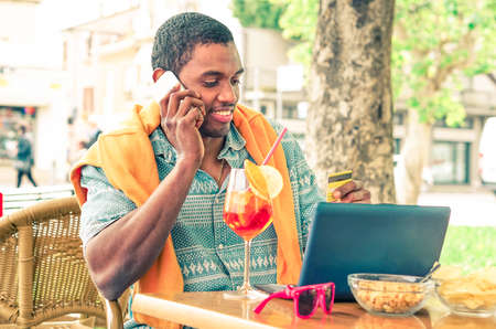 African american man using credit card with mobile phone and laptop internet connected - Young black male talking at cellphone sitting in cafe bar with drink on table - Desaturated vintage filter lookの写真素材