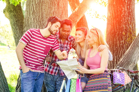 Group of multiracial friends looking map at sunset in city park - Cheerful students having fun in a bike tour searching the way outdoor - Concept of tourism and ecological transport focus on left guyの写真素材