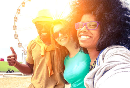 Afro hair girl taking selfie with best friends at ferris wheel at sunset - Happy multiracial group of students having fun with smartphone photo camera outside- Warm sun halo filter with vintage tonesの写真素材
