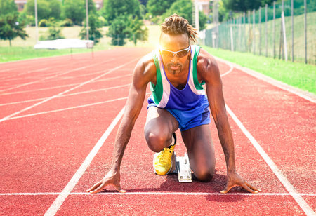 Afro american athlete ready to sprint at starting line - Runner kneeling on blocks of red athletics track - Concept of human concentration at sport competition - Soft vintage filter and sun reflectionの写真素材