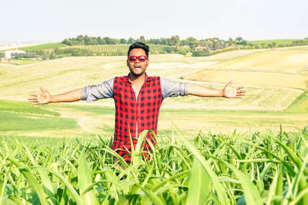 Young indian farmer with open arms in a corn field - Cheerful asian man walking through countryside with a freedom attitude and positive expression - Concept of life joyful moment and human happinessの写真素材