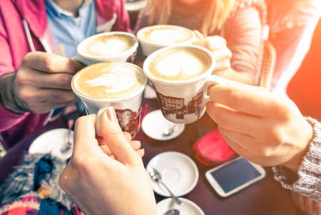 Group of friends cheers with cappuccino cup in cafe bar with phone on table - Family having fun drinking together at restauran in winter season closeup scene with soft vintage filter and sun haloの写真素材