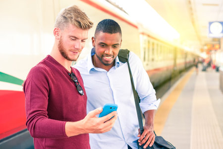 Interracial couple of men looking mobile phone screen at train station - Handsome guys commuters with casual clothing using smartphone at railway platform - Concept of everyday urban  lifestyleの写真素材