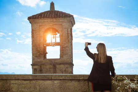Silhouette of woman taking photo with smart phone of old bell tower at sunset - Back view of female tourist shooting church steeple - Concept of tourism and technology - Backlit image soft vignettingの写真素材