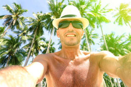 Cheerful man selfie with palm trees background in tropical island - Self male tourist portrait introspection photo - Concept of single trip holidays and happiness - Soft focus due sun flare effectの写真素材