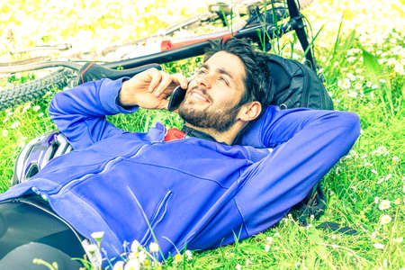 Young biker man lying on green grass talking on mobile - Handsome happy guy relaxing at park next to sport bike is using phone and looking up - Concept of healthy human activity in spring and summerの写真素材