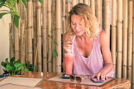 Young traveler woman drinking fruit cocktail in tropical beach bar - Girl sitting in cafe smiles holding straw and texting message with mobile phone - Concept of  relaxing holiday around the worldの写真素材