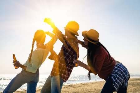 Cheering friends party and selfie holding beer bottle at sunset beach festival - Silhouette of happy teenagers having fun taking photo - University friendship concept in backlight scene with sun flareの写真素材