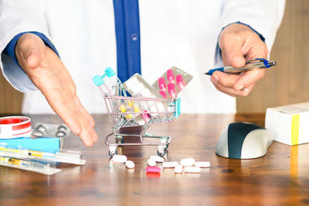 Pills and medicinal products inside shopping trolley - Doctor hands showing tablets and pharmaceutical package on wooden table - Concept of online medicines purchase risk - Focus on little cartの写真素材