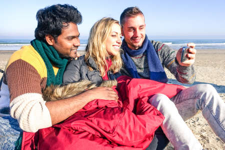 Multiracial young friends taking selfie on the beach in sunny winter day - Multiethnic teenagers having fun looking mobile phone - Concept of friendship happy moment with colorful vintage filter tonesの写真素材