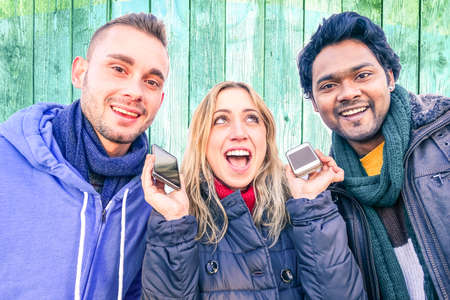 Surprised woman holding mobile smart phones and amused multiracial friends on wooden wall background - Funny girl between different guys - Concept of human astonishment in everyday lifeの写真素材