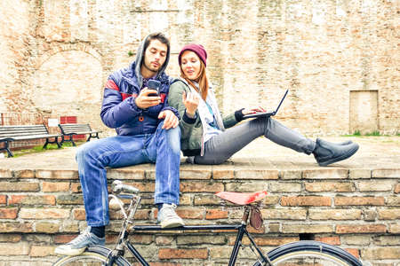 Teenage couple using phone and notebook outdoor at winter time - Best friends looking internet content sitting on old urban wall  - Concept of young people lifestyle with modern technologies and trendの写真素材