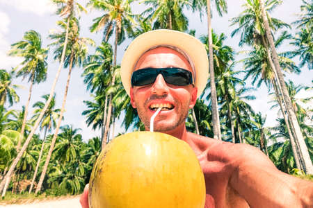Cheerful man beach selfie drinking coconut milk with palm trees background at tropical island - Middle age male tourist taking self photo on sunny day - Concept of single trip holidays and happinessの写真素材