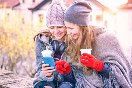 Young women looking phone smiling and holding coffee cup outdoor - Happy best girlfriends pointing mobile screen sitting in city park on winter holiday at sunset - Focus on right female warm filterの写真素材
