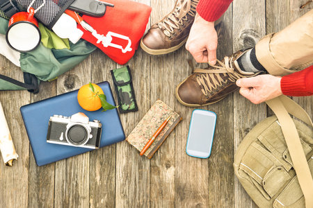 Travel man tying boots for trekking day top view of items for hiking and adventure - Active male hands on shoelaces above angle with camera mobile phone and backpack on old wooden floor backgroundの写真素材